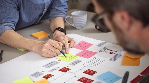 To mænd arbejder sammen om et projekt, mens de skriver på farvede post-it sedler og en stor plakat med diagrammer og information. En kaffekop kan ses i baggrunden, hvilket skaber en kreativ og samarbejdende atmosfære.